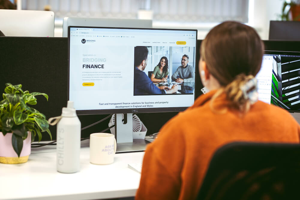 Web designer Alice in orange shirt reviewing finance website project on desktop monitor in modern office workspace with plants and professional setup