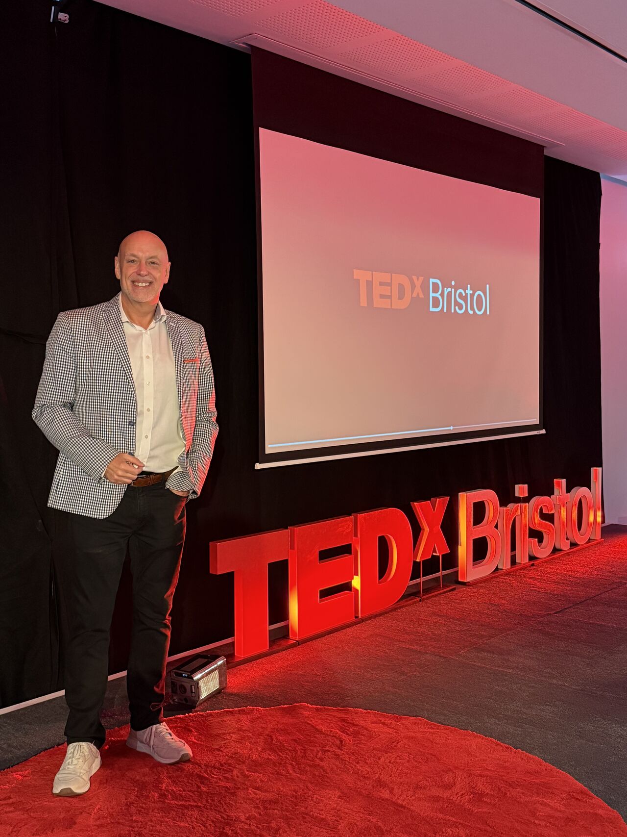 Andrew Scott standing on the red TEDx stage at TEDxBristol, smiling at the camera with the event logo displayed on the screen behind him.